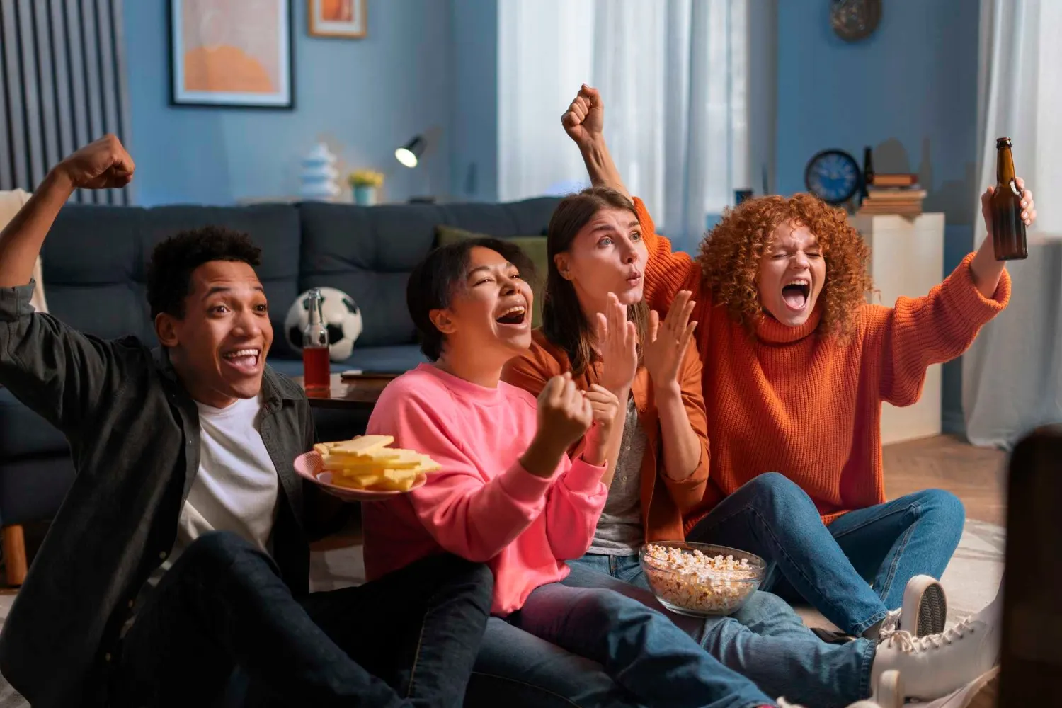 Vier vrienden juichen enthousiast op de bank terwijl ze samen live voetbal kijken met chips, popcorn en bier